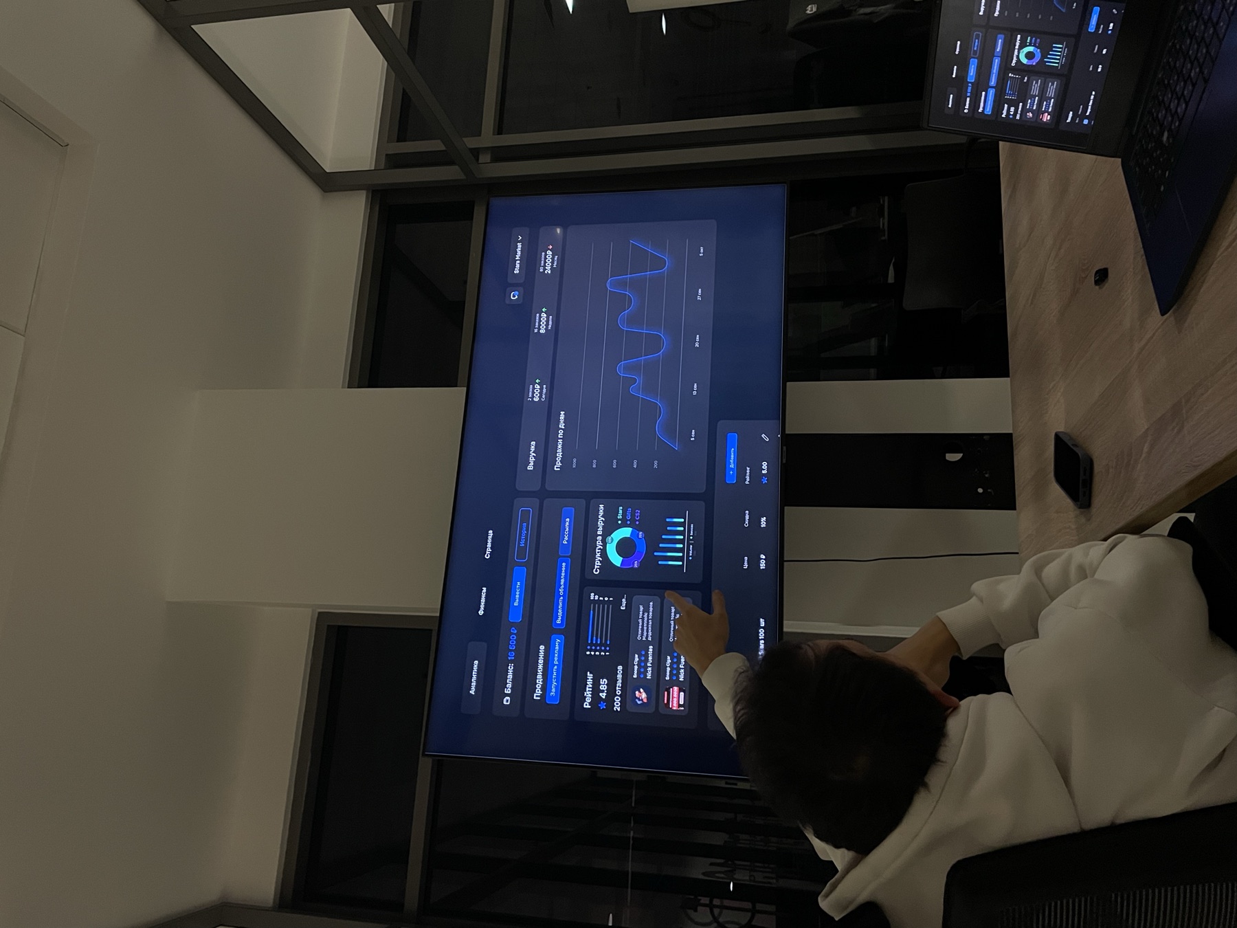 A sales manager pointing at a ranked list on a wall screen while a colleague works through it on a laptop